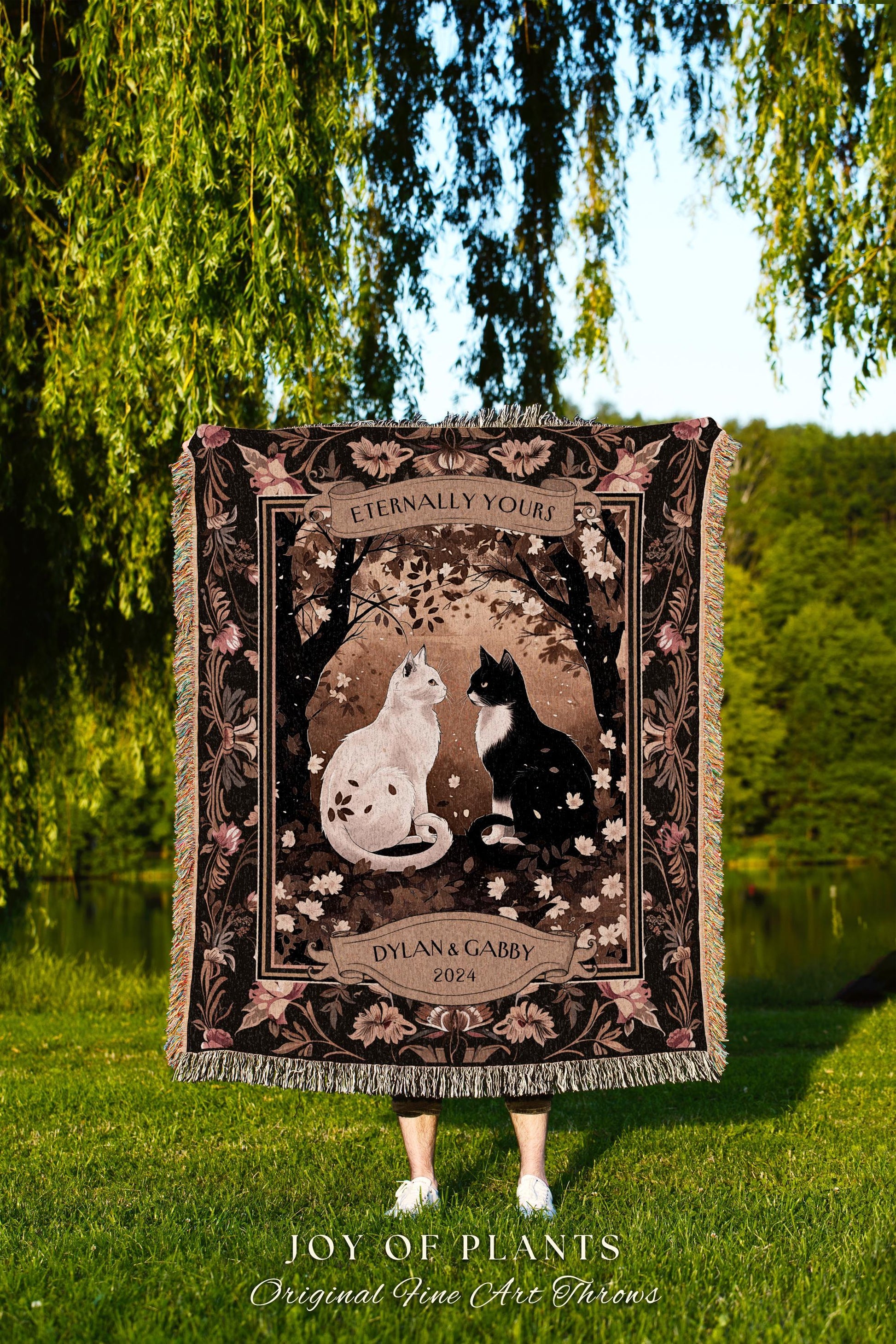 May include: A tapestry throw blanket with a black background and a brown and white floral pattern. The center of the blanket features a white cat and a black cat sitting facing each other. The blanket has a banner at the top that reads 'Eternally Yours' and a banner at the bottom that reads 'Dylan & Gabby 2024.' The blanket is displayed in front of a green grassy area and a body of water.