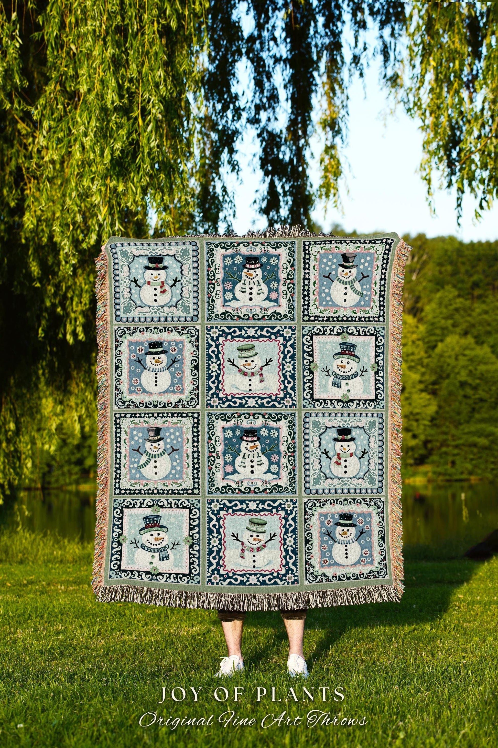 May include: A cozy woven throw blanket features a patchwork design of 12 framed snowman illustrations. Each square shows a unique snowman in varying poses and hats against a muted blue and green background. The blanket has fringed edges and is made of a soft, textured fabric. The text 'JOY OF PLANTS Original Fine Art Throws' is displayed at the bottom.