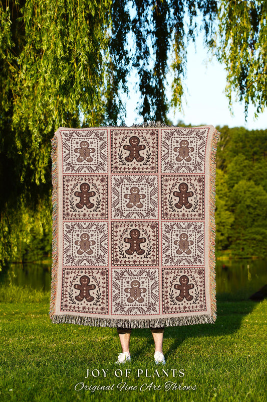 May include: A woven throw blanket with a brown gingerbread man pattern on a cream background. The blanket has a fringe border. The text 'JOY OF PLANTS Original Fine Art Throws' is at the bottom of the image.