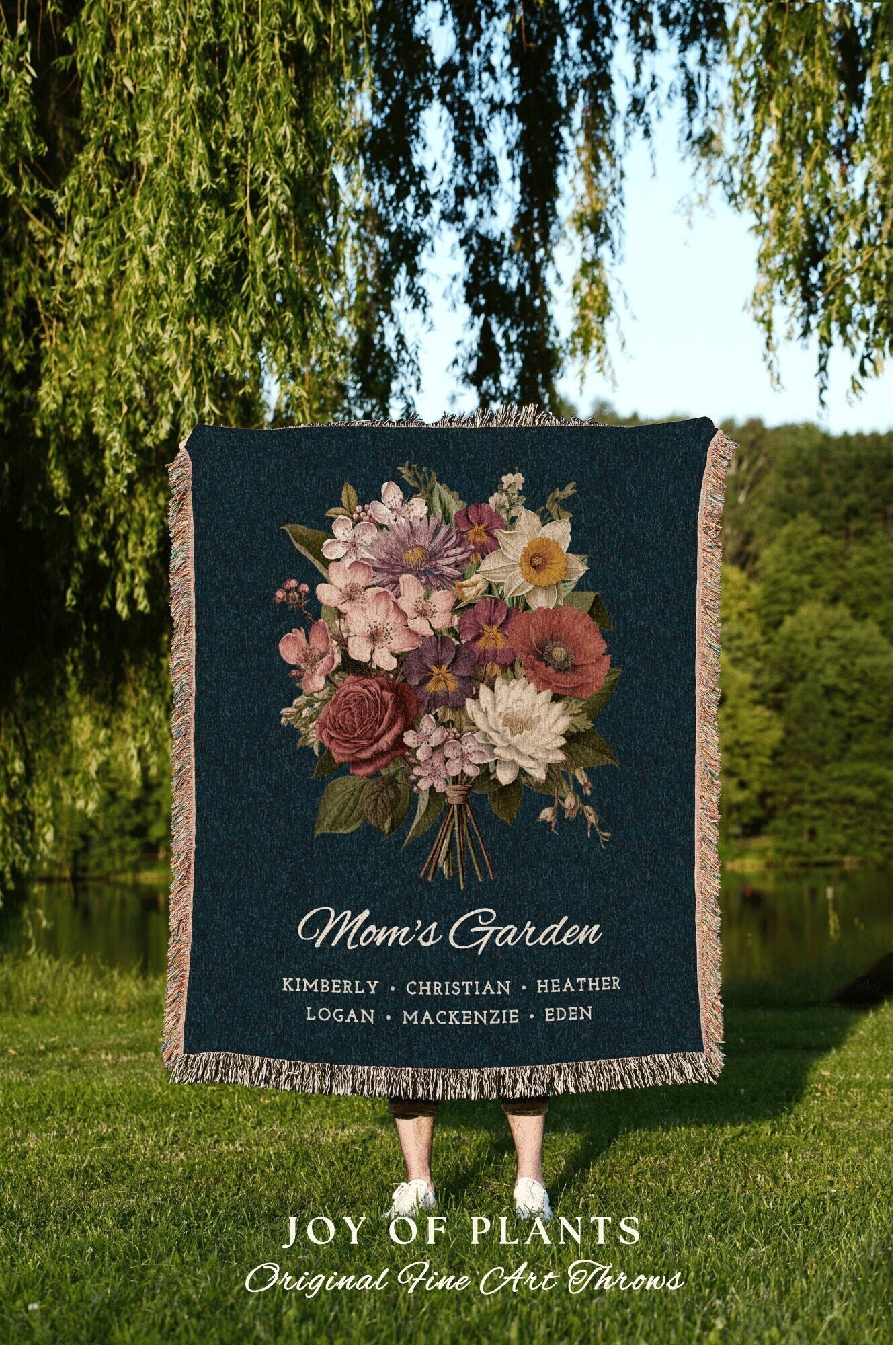 May include: A woven tapestry throw blanket with a dark blue background and a colorful floral bouquet in the center. The text 'Mom's Garden' is below the bouquet, followed by a list of names: Kimberly, Christian, Heather, Logan, Mackenzie, Eden. The text 'JOY OF PLANTS Original Fine Art Throws' is at the bottom of the blanket.