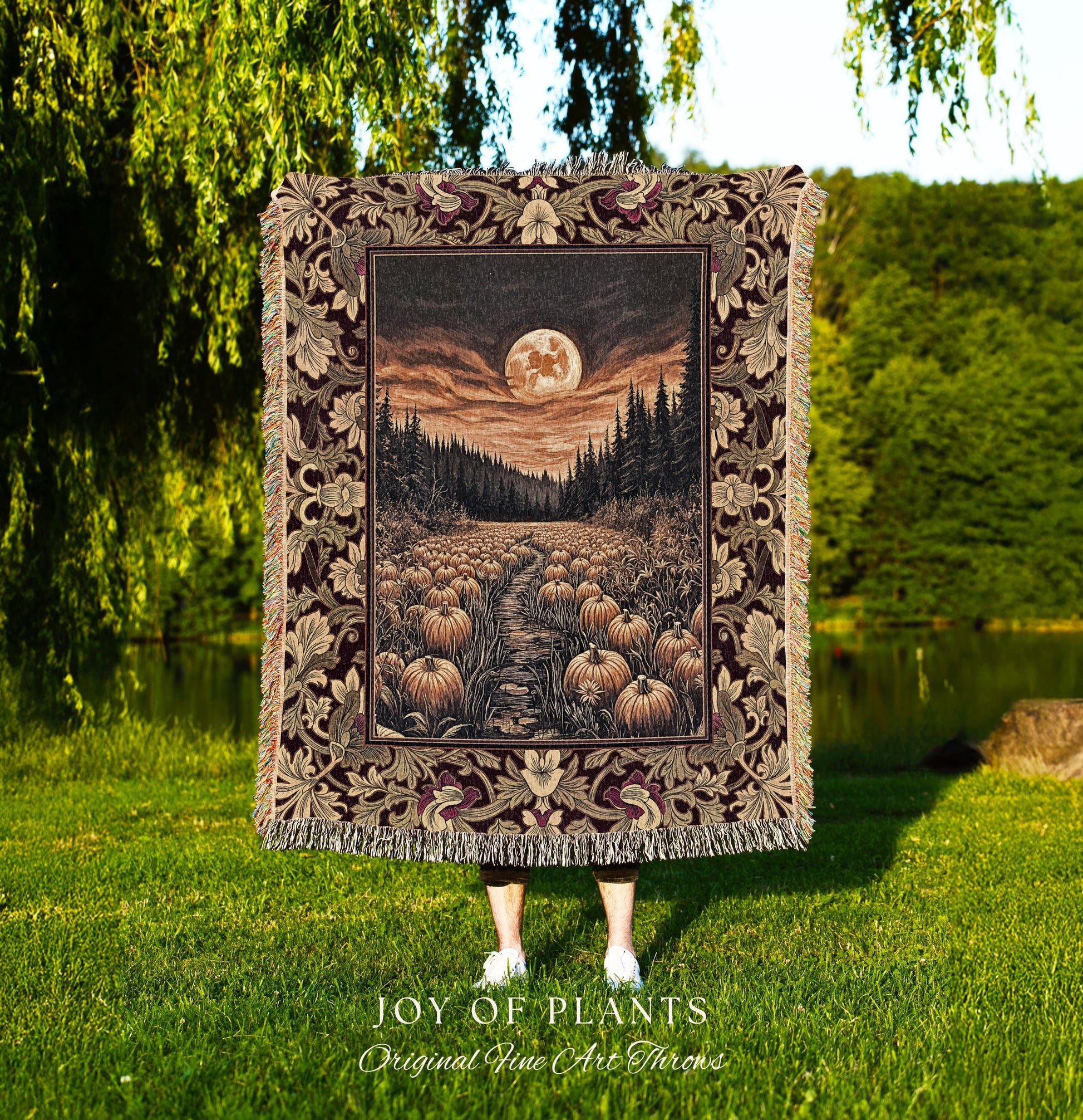 May include: A tapestry throw blanket featuring a nighttime landscape with a full moon, a path lined with pumpkins, and a forest. The border has a floral design with burgundy and tan colors. The blanket is held up in front of a person standing in a grassy area. The text 'JOY OF PLANTS Original Fine Art Throws' is at the bottom.