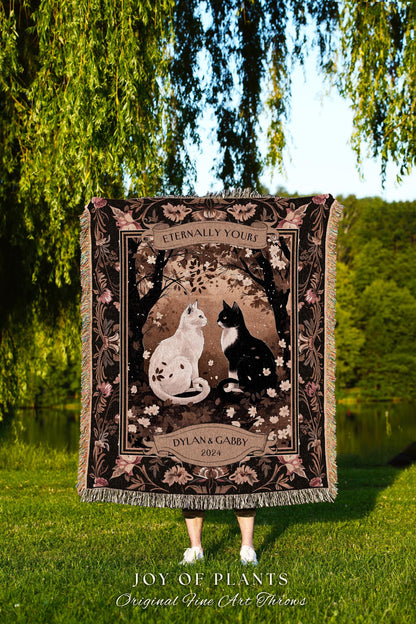 May include: A tapestry throw blanket with a black background and a brown and white floral pattern. The center of the blanket features a white cat and a black cat sitting facing each other. The blanket has a banner at the top that reads &#39;Eternally Yours&#39; and a banner at the bottom that reads &#39;Dylan & Gabby 2024.&#39; The blanket is displayed in front of a green grassy area and a body of water.