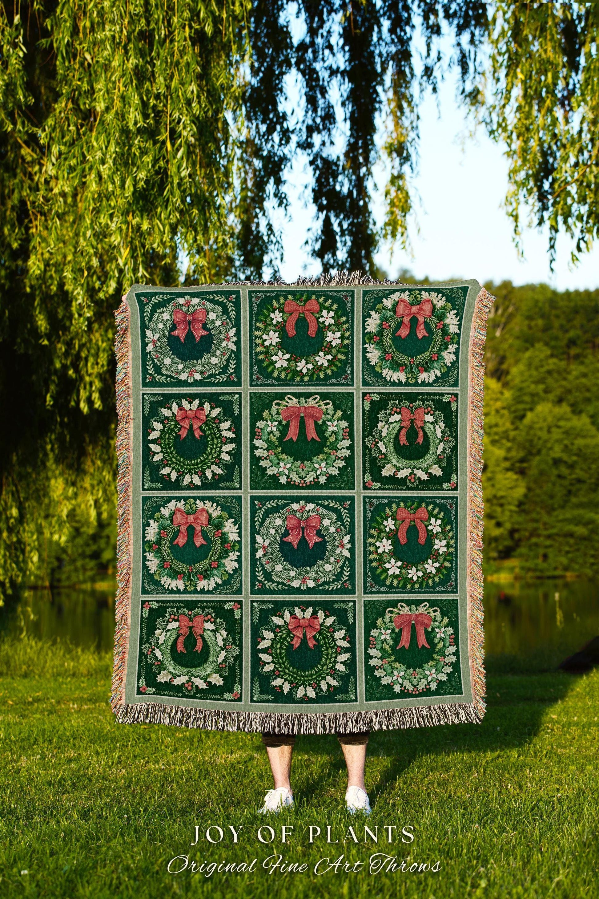 May include: A green woven throw blanket with a repeating pattern of wreaths with red bows. The throw blanket has fringe on the edges. The text &#39;JOY OF PLANTS Original Fine Art Throws&#39; is printed at the bottom of the image.