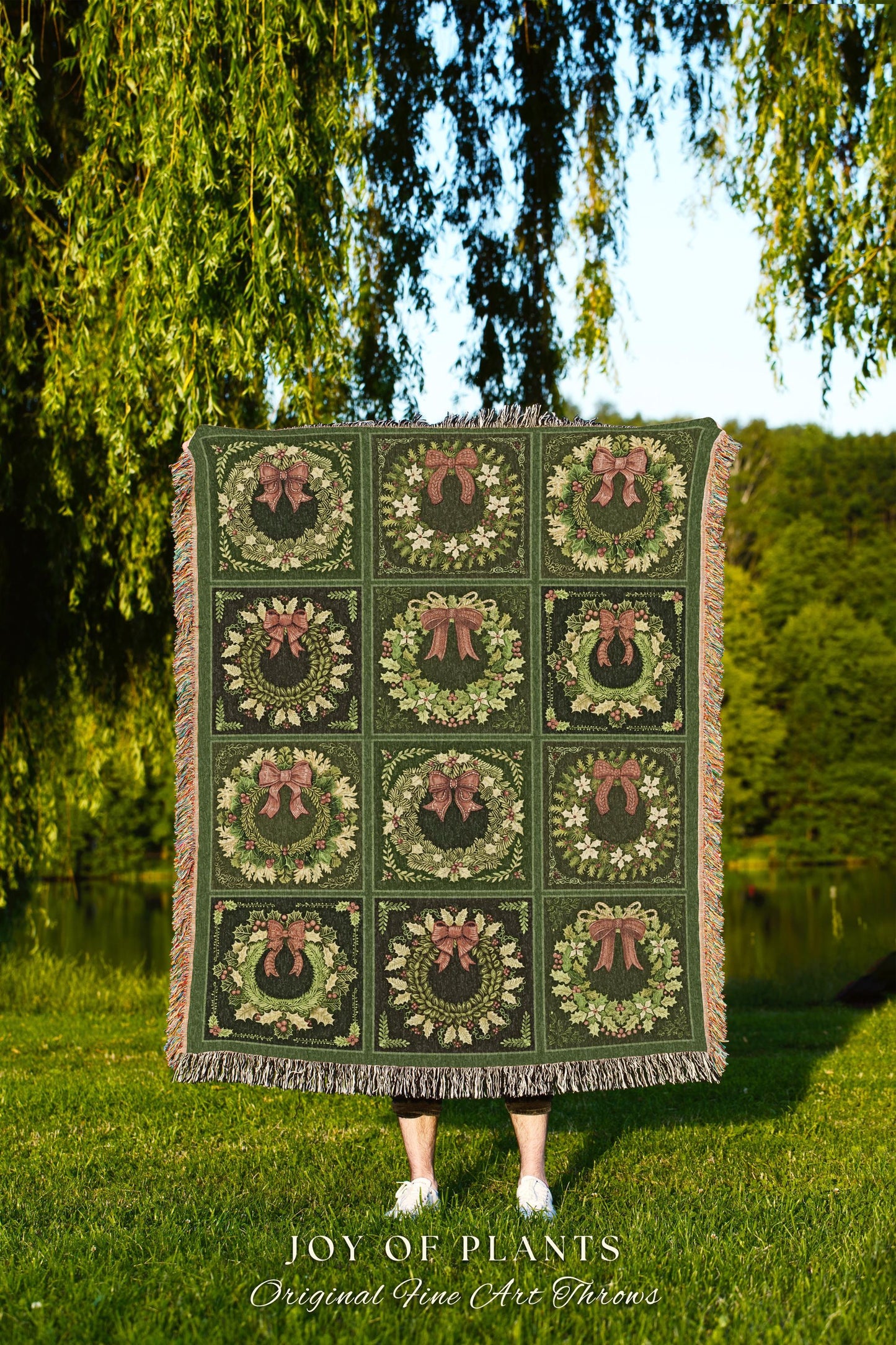 May include: A green woven throw blanket with a fringed border. The blanket features a repeating pattern of nine green wreaths with red bows. The text &#39;JOY OF PLANTS Original Fine Art Throws&#39; is printed below the blanket.