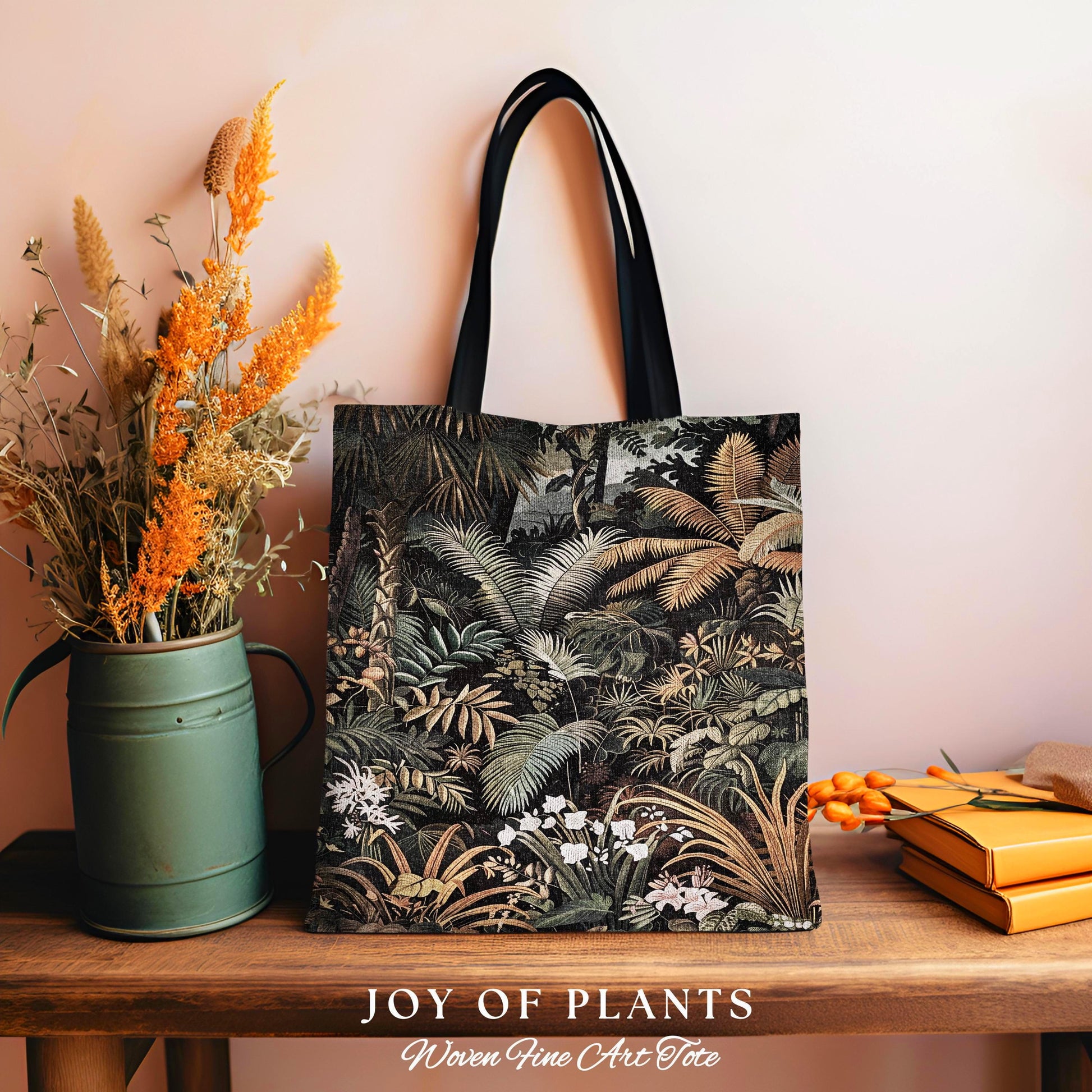 May include: A tote bag with a jungle print design. The bag features a black handle and a detailed illustration of various plants and foliage in shades of green, brown, and white. The words &#39;JOY OF PLANTS&#39; and &#39;Woven Fine Art Tote&#39; are printed at the bottom.