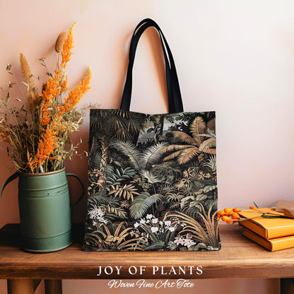 May include: A tote bag with a jungle print design. The bag features a black handle and a detailed illustration of various plants and foliage in shades of green, brown, and white. The words &#39;JOY OF PLANTS&#39; and &#39;Woven Fine Art Tote&#39; are printed at the bottom.