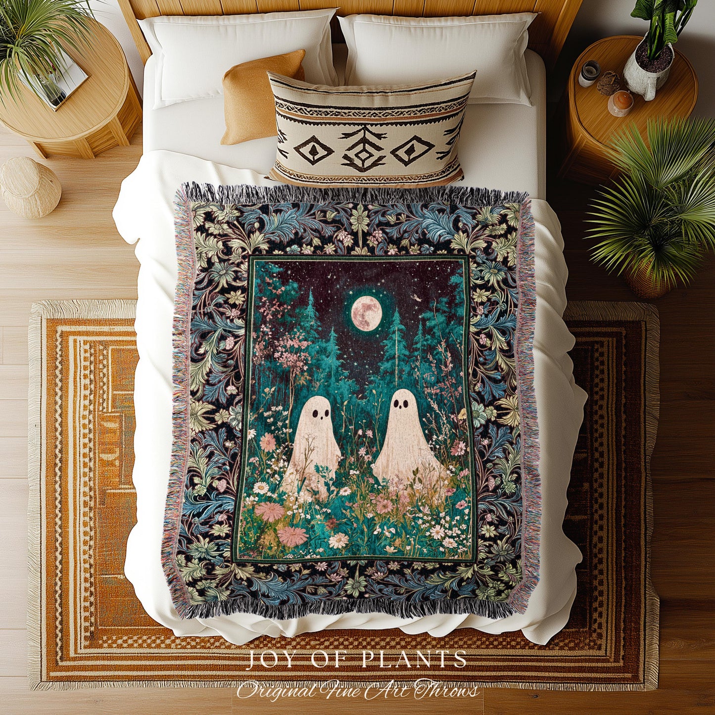May include: A decorative throw blanket with a whimsical design. The blanket features a dark teal border with floral patterns. The center showcases a night scene with two ghost figures in a field of flowers under a full moon. The blanket is placed on a bed with white bedding, and a patterned pillow is visible.