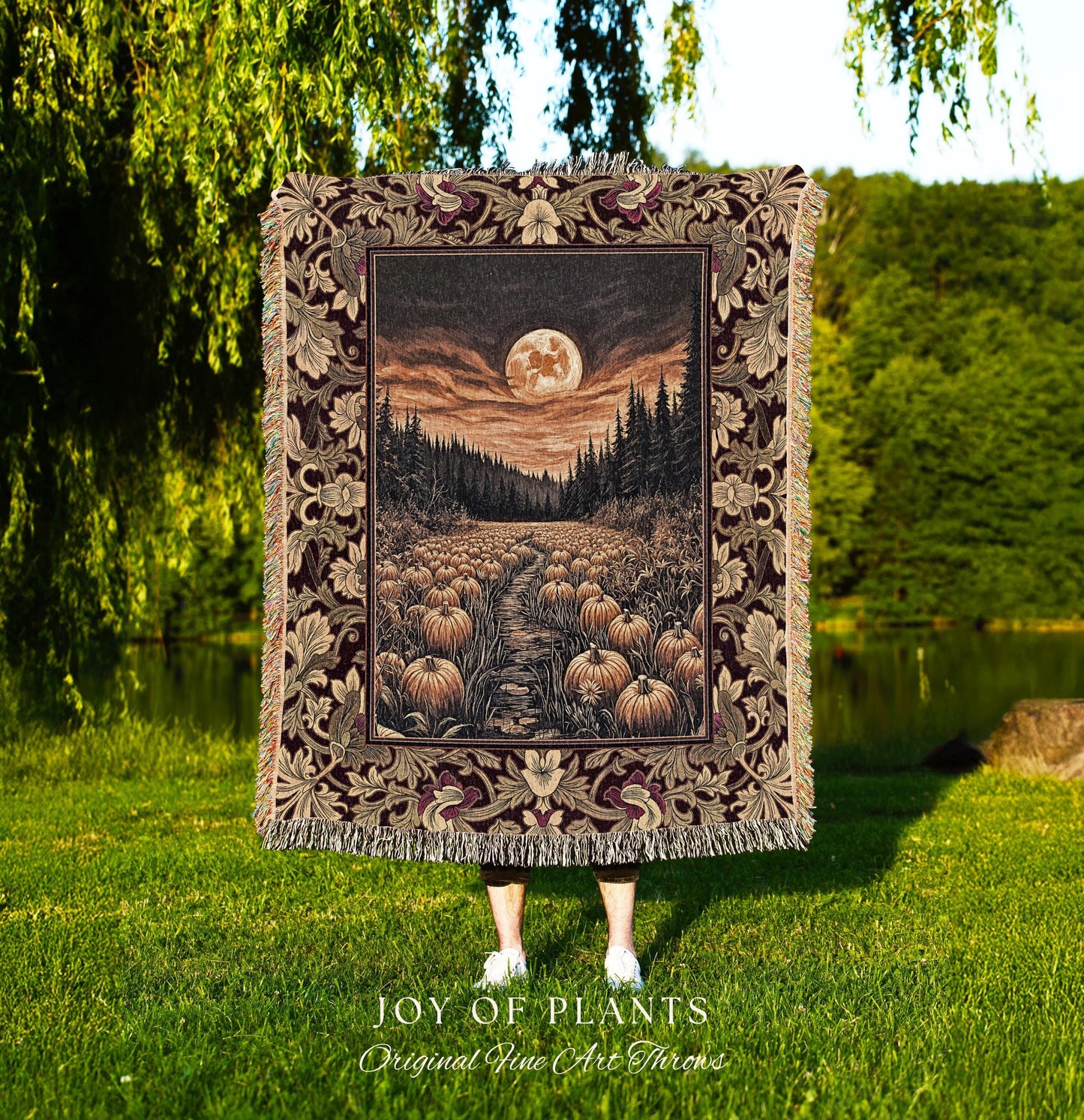 May include: A tapestry throw blanket featuring a nighttime landscape with a full moon, a path lined with pumpkins, and a forest. The border has a floral design with burgundy and tan colors. The blanket is held up in front of a person standing in a grassy area. The text &#39;JOY OF PLANTS Original Fine Art Throws&#39; is at the bottom.