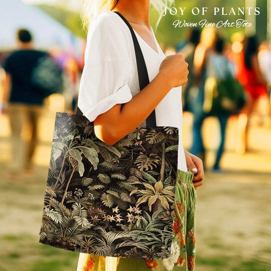May include: A tote bag with a dark green and black jungle print. The bag features a detailed pattern of tropical plants and foliage. The bag is carried by a person wearing a white top and green skirt. Text on the bag reads &#39;JOY OF PLANTS Woven Fine Art Tote&#39;.