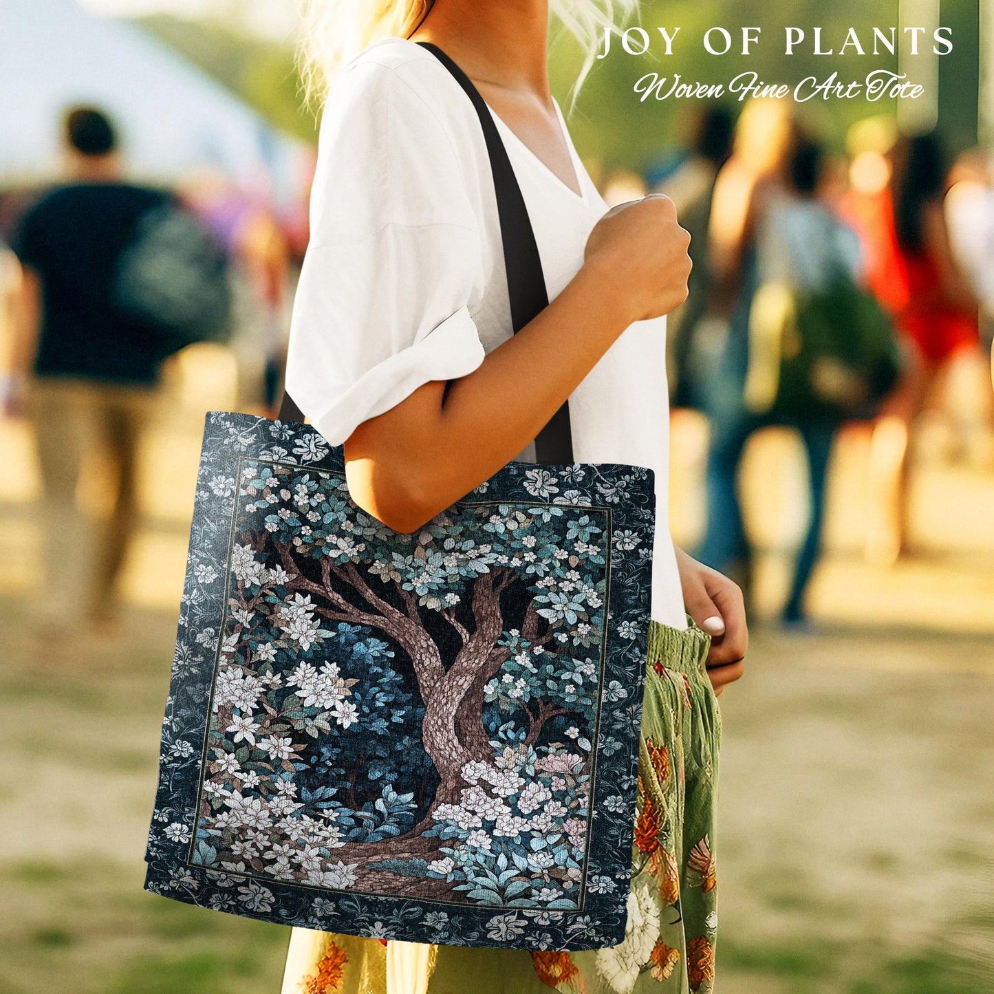 May include: A tote bag featuring a detailed illustration of a tree with white flowers and blue foliage. The bag has a dark blue border and a black strap. The text "JOY OF PLANTS Woven Fine Art Tote" is at the top.