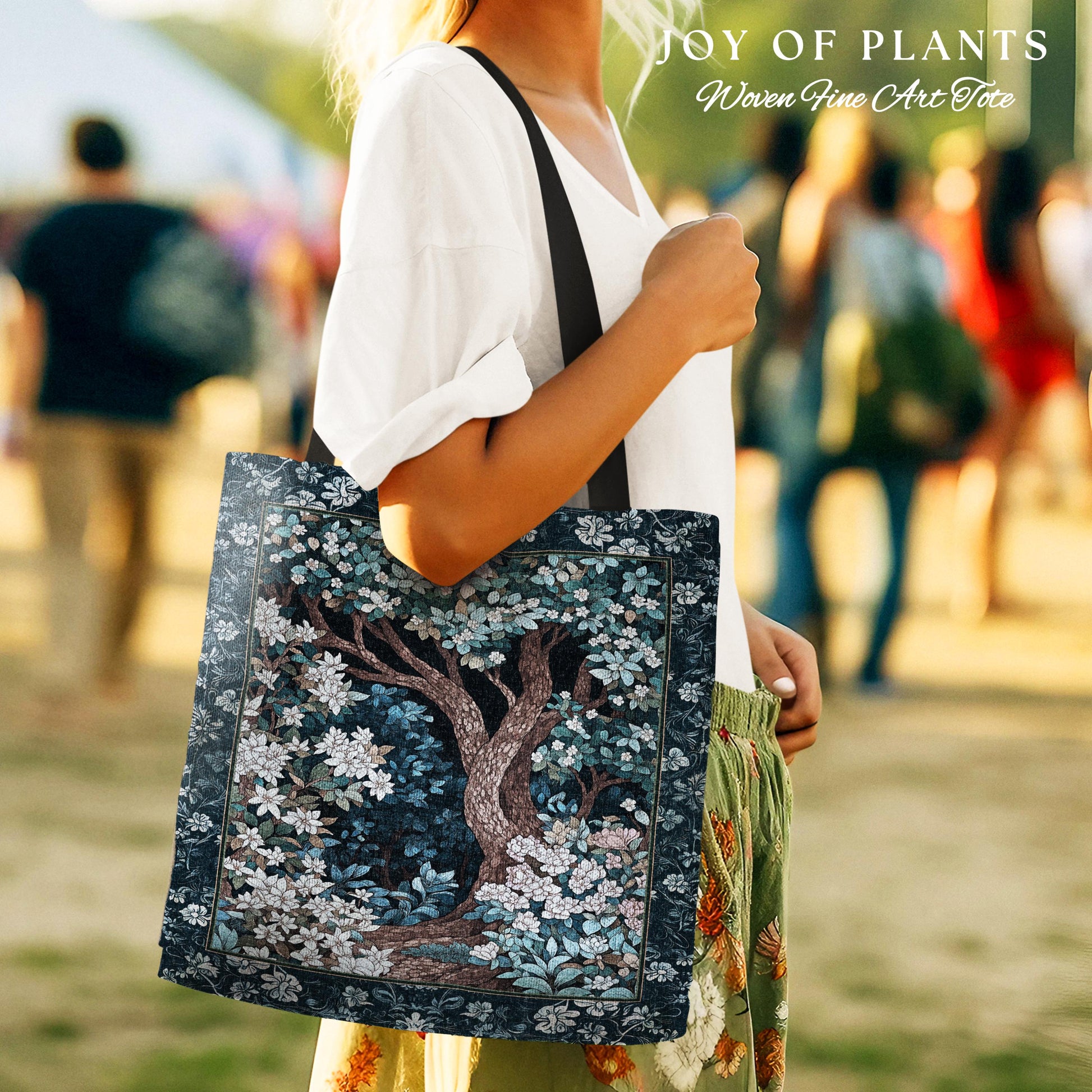May include: A tote bag featuring a detailed illustration of a tree with white flowers and blue foliage. The bag has a dark blue border and a black strap. The text "JOY OF PLANTS Woven Fine Art Tote" is at the top.