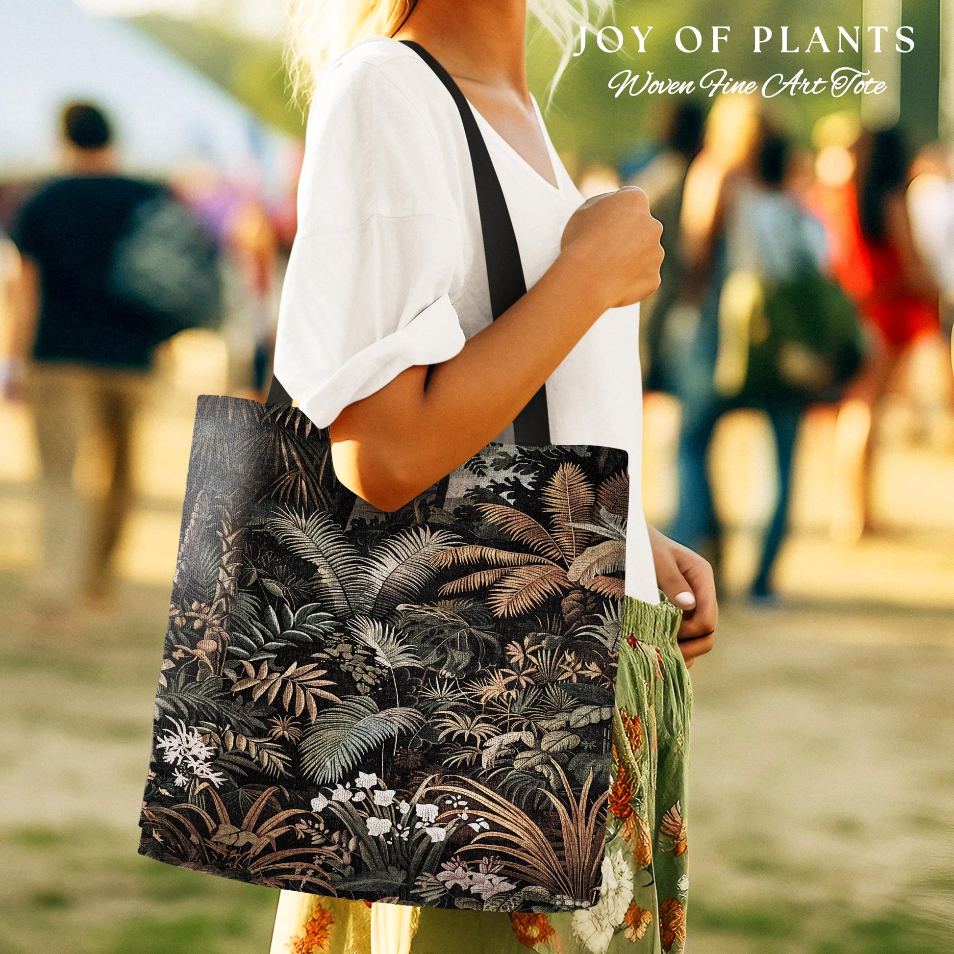 May include: A black tote bag with a detailed botanical print featuring various plants and flowers. The bag has a black strap and the text "JOY OF PLANTS Woven Fine Art Tote" is visible in the upper right corner. The bag is carried by a person.
