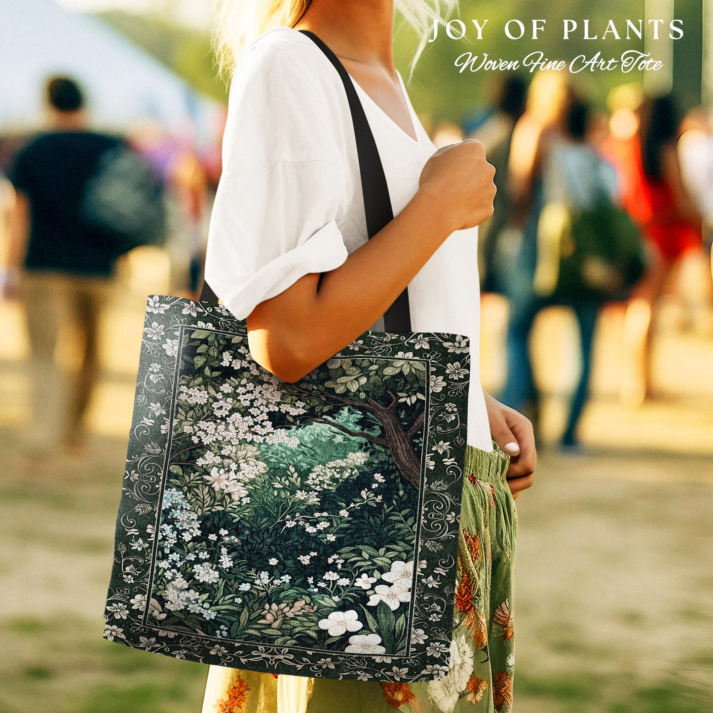 May include: A tote bag with a dark green floral design featuring white flowers and a tree. The bag has a black strap and the text "JOY OF PLANTS Woven Fine Art Tote" at the top. The bag is being held by a person wearing a white shirt and green pants.