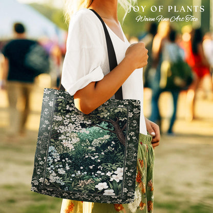 May include: A tote bag with a dark green floral design featuring white flowers and a tree. The bag has a black strap and the text "JOY OF PLANTS Woven Fine Art Tote" at the top. The bag is being held by a person wearing a white shirt and green pants.