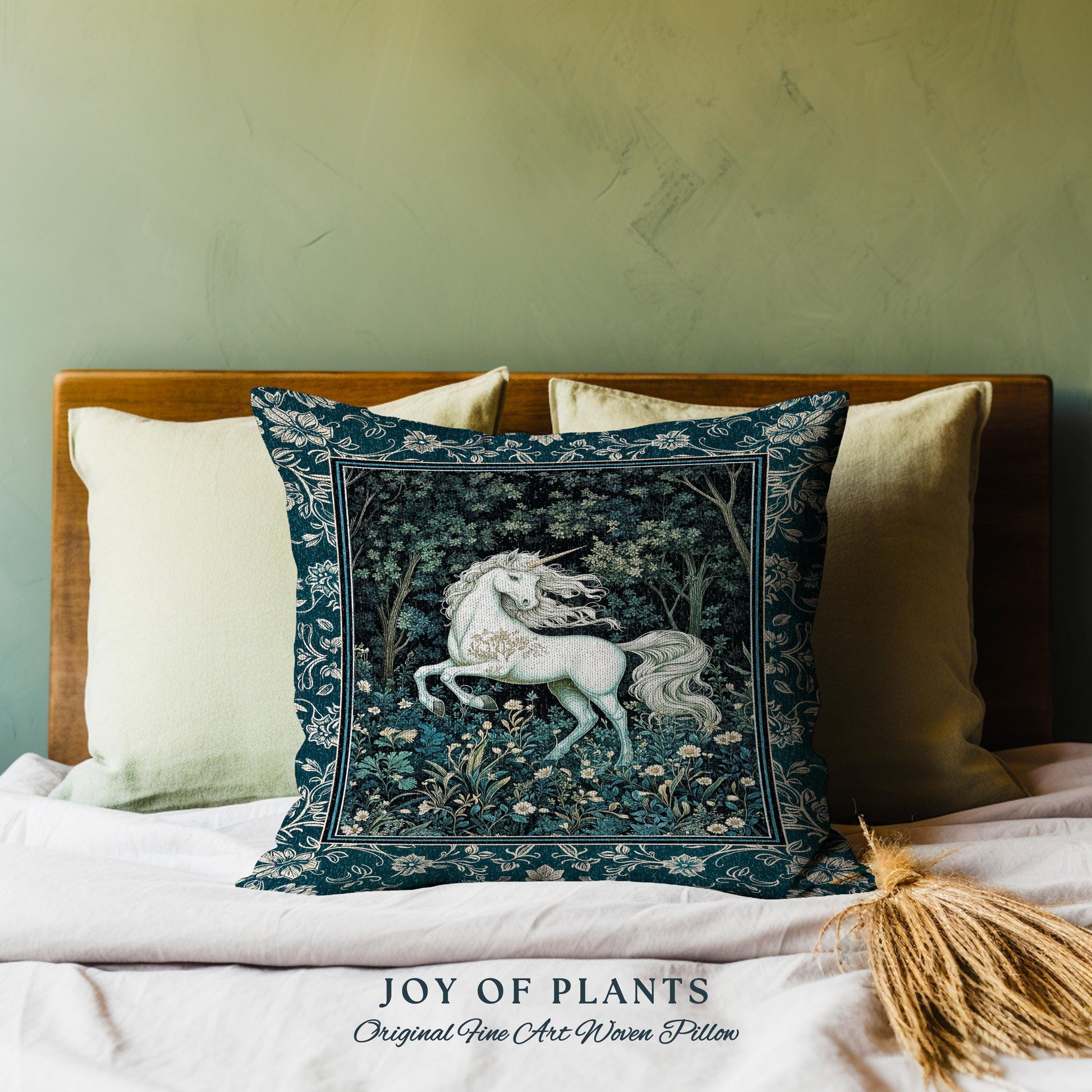 May include: A decorative square pillow with a dark teal border and a central image of a white unicorn in a forest setting. The pillow features the text "JOY OF PLANTS Original Fine Art Woven Pillow" at the bottom. The pillow is placed on a bed with other pillows.