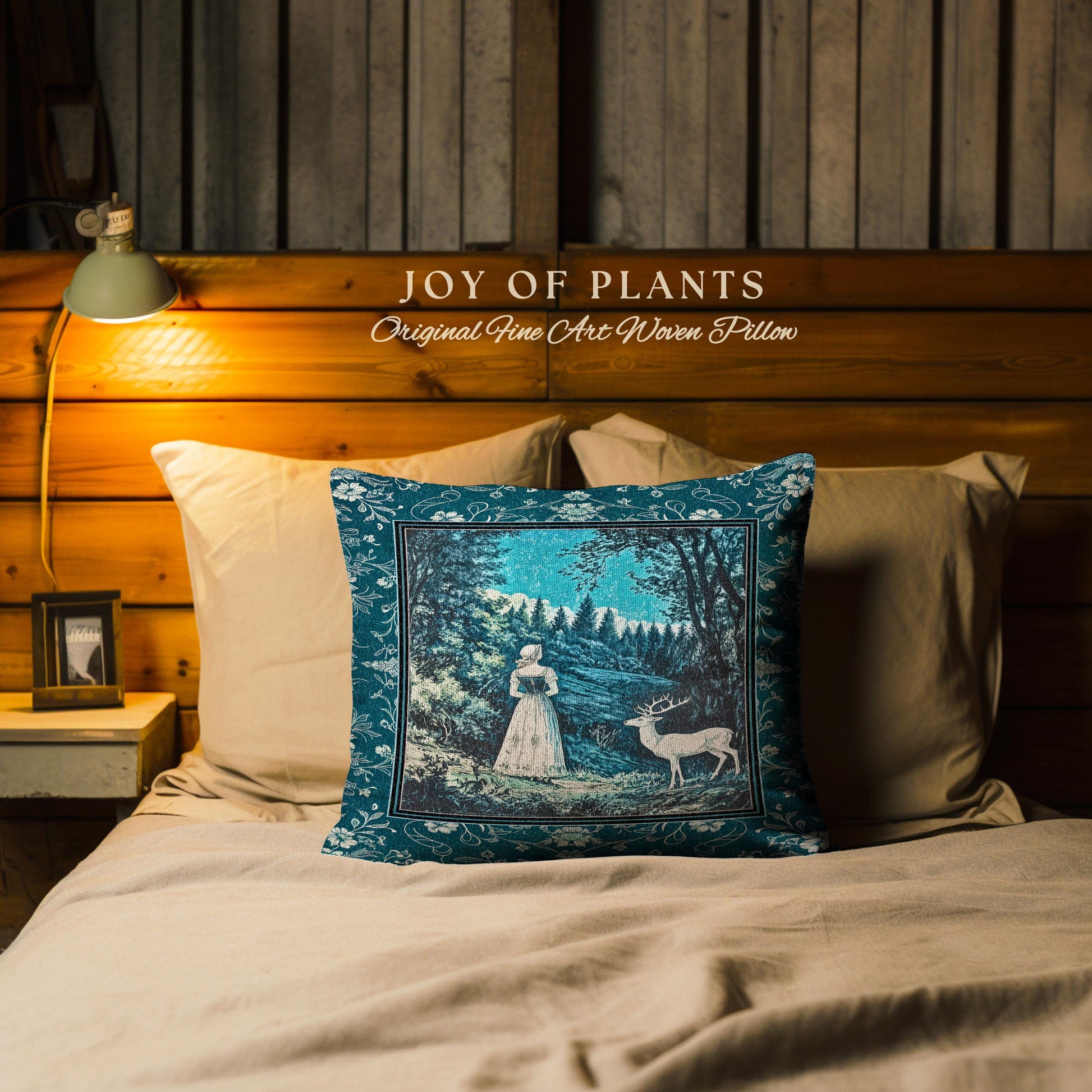 May include: A decorative pillow with a teal border and a central image of a woman and a deer in a forest scene. The pillow is on a bed with other pillows. The text "JOY OF PLANTS Original Fine Art Woven Pillow" is above the pillow.
