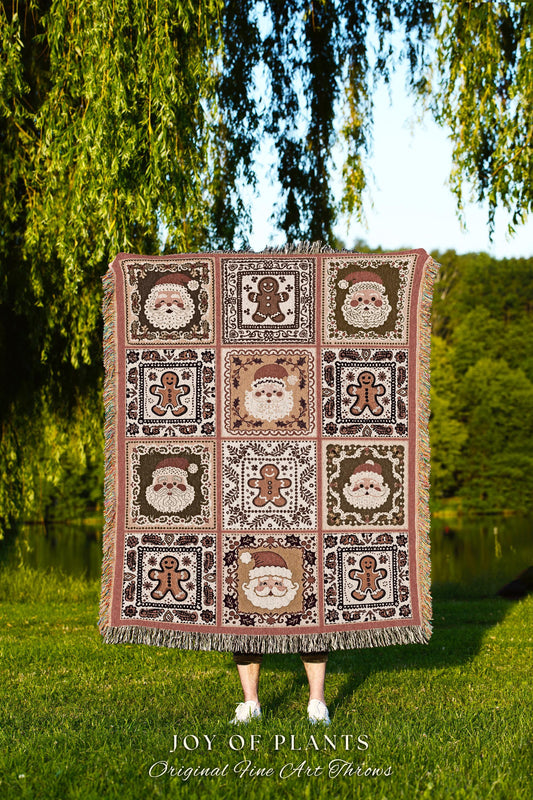 May include: A Christmas-themed woven throw blanket features a patchwork design with repeating patterns of Santa Claus and gingerbread men in shades of brown, green, and beige against a blush pink background. The blanket has fringed edges and is displayed against a blurred outdoor backdrop.  The text 'JOY OF PLANTS Original Fine Art Throws' is visible at the bottom.