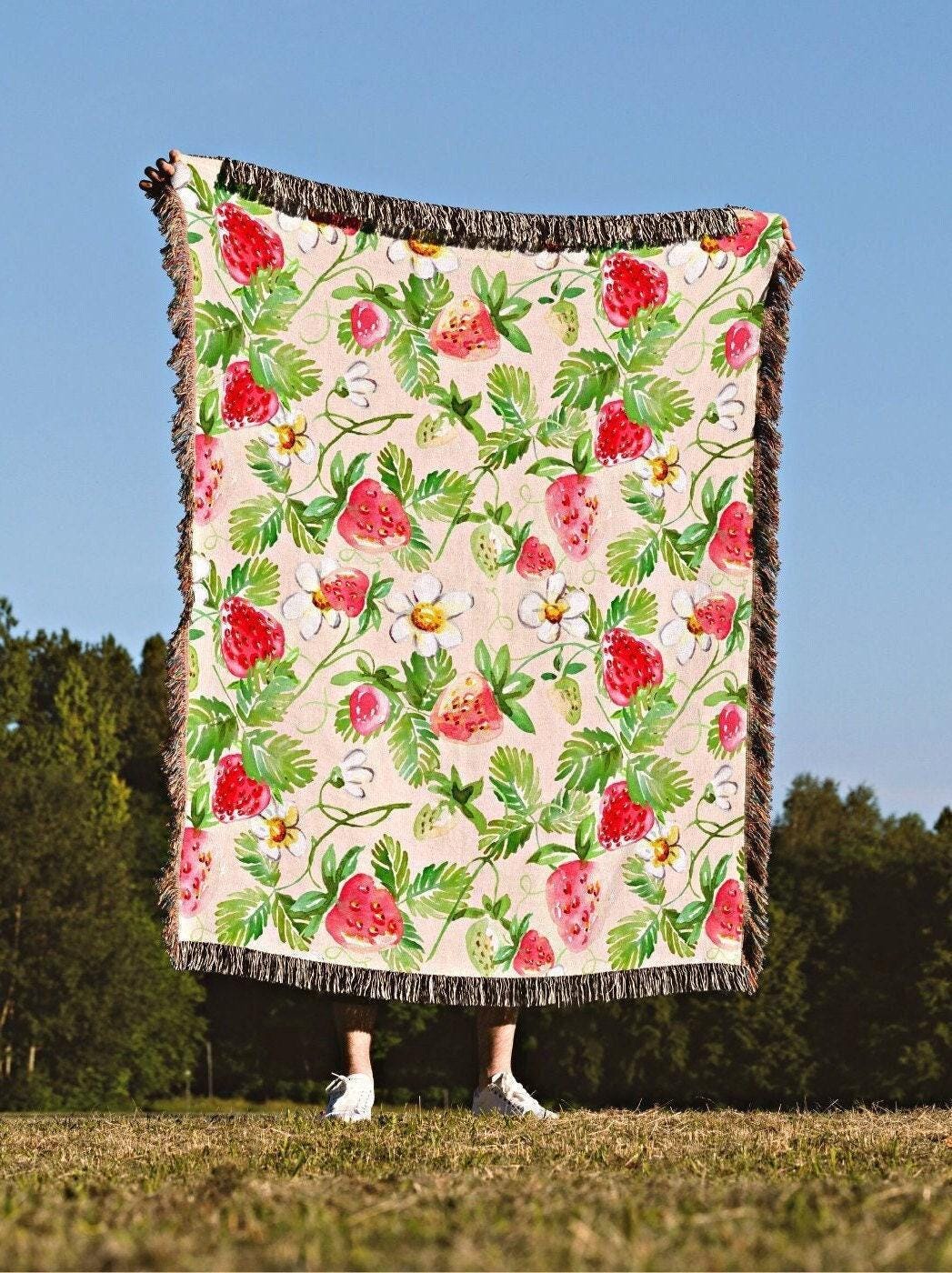 May include: A rectangular throw blanket with a repeating pattern of red strawberries, green leaves, and white flowers on a light pink background. The blanket has a fringed edge and is held up against a blue sky.