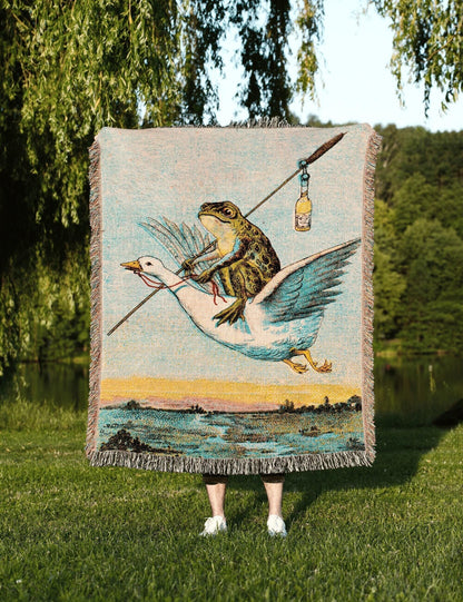 May include: A woven tapestry throw blanket featuring a whimsical scene of a frog riding a goose, holding a fishing rod with a bottle. The blanket has a fringed edge and a landscape background with a lake and trees. The color palette includes blue, green, yellow, and beige.