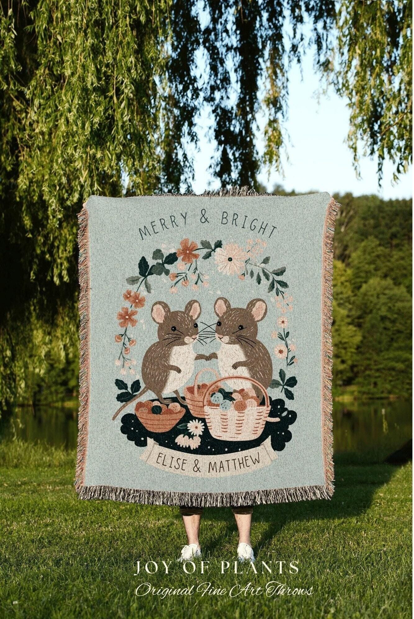 May include: A light blue throw blanket with a whimsical design featuring two brown mice holding hands, surrounded by floral elements and baskets. The text "MERRY & BRIGHT" and "ELISE & MATTHEW" are displayed on the blanket.
