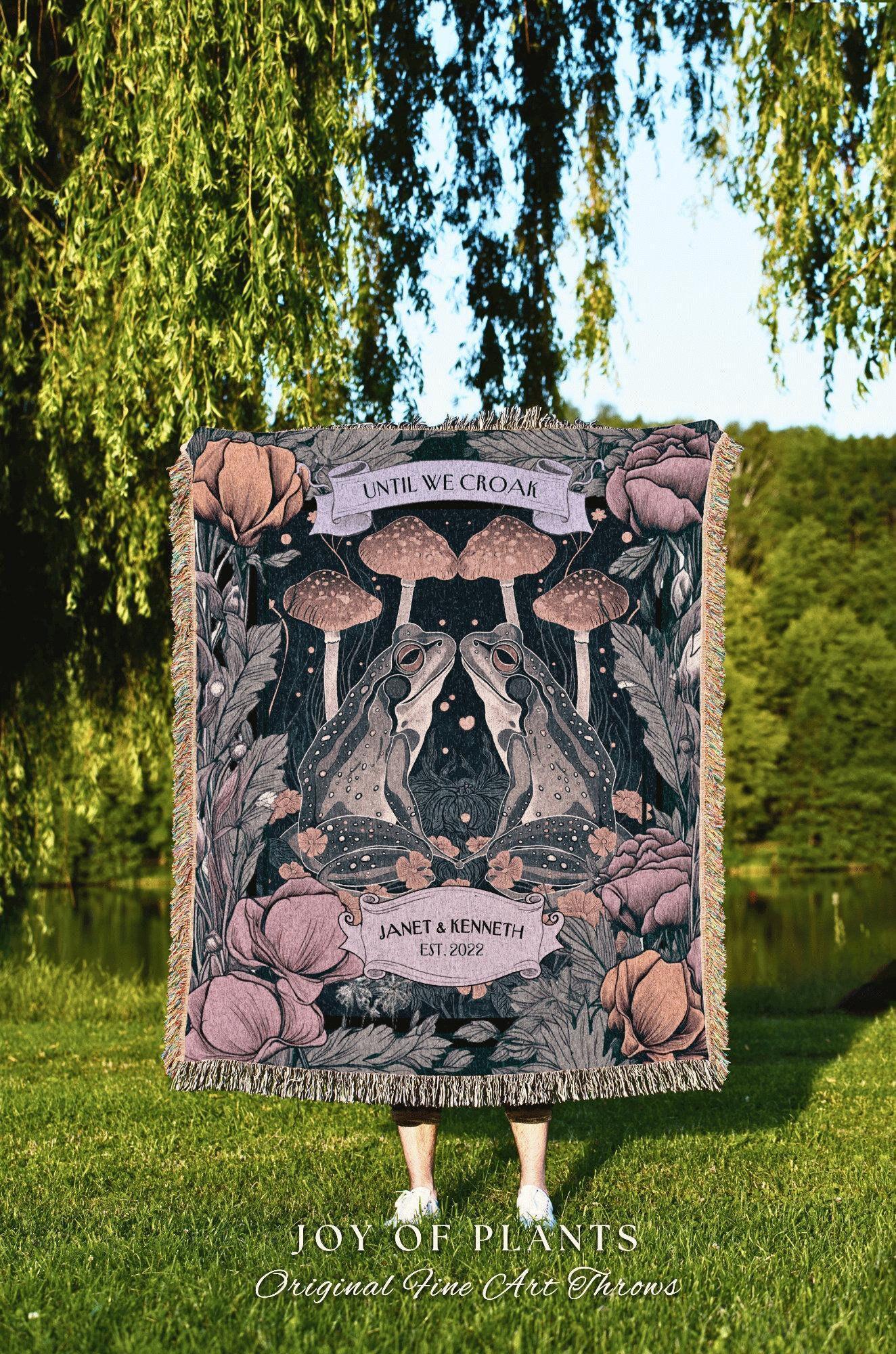 May include: A woven throw blanket featuring a whimsical design with two facing frogs, mushrooms, and floral elements in shades of pink, green, and black. The blanket includes the text "UNTIL WE CROAK" and "JANET & KENNETH EST. 2022".