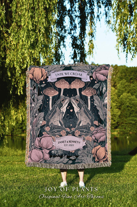 May include: A woven throw blanket featuring a whimsical design with two facing frogs, mushrooms, and floral elements in shades of pink, green, and black. The blanket includes the text "UNTIL WE CROAK" and "JANET & KENNETH EST. 2022".