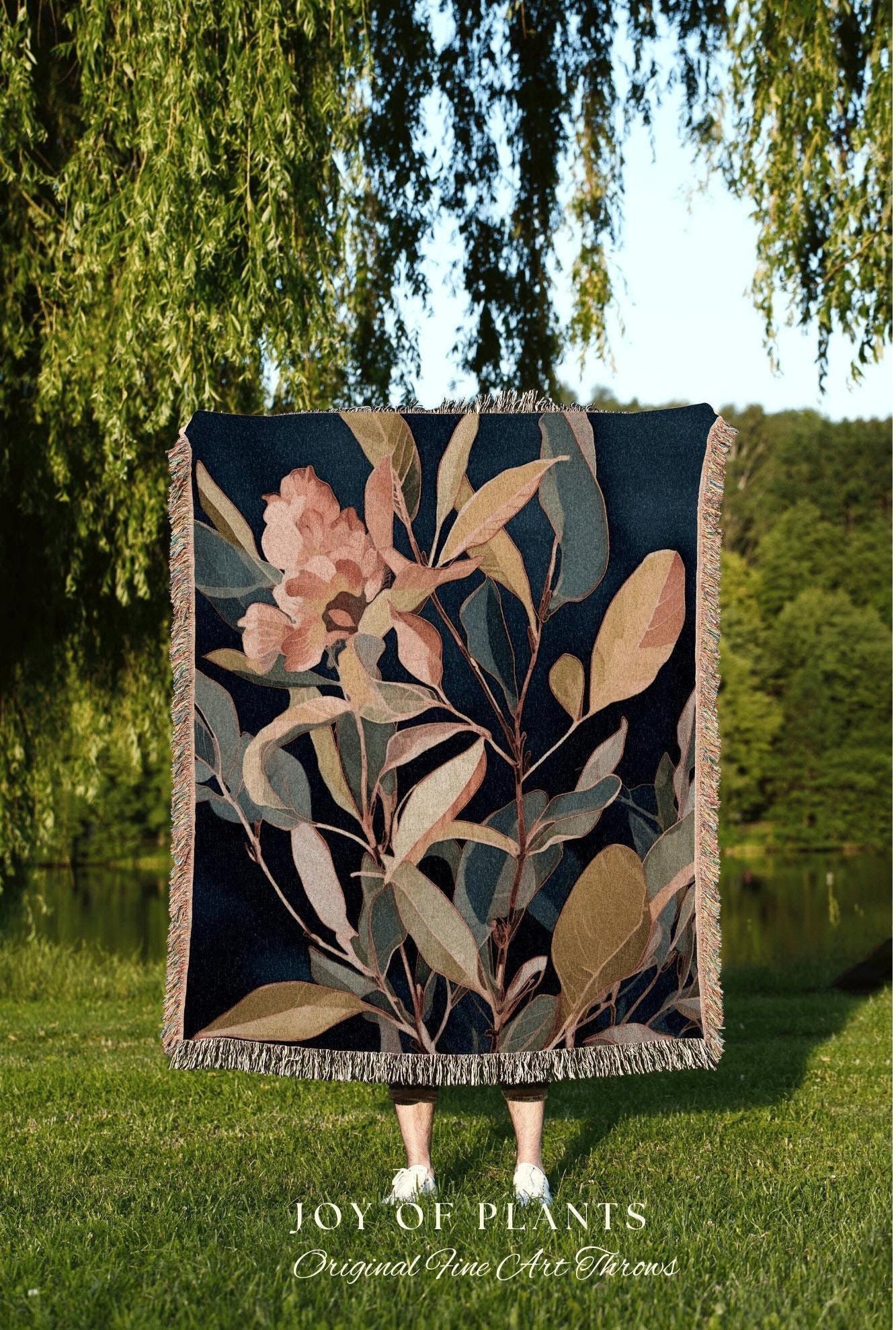 May include: A dark blue throw blanket with a botanical design featuring peach and green leaves and flowers. The blanket has a fringed edge and the text "JOY OF PLANTS Original Fine Art Throws" at the bottom.
