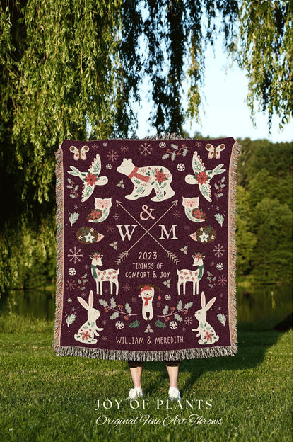 May include: A woven tapestry throw blanket with a burgundy background and a festive winter scene. The blanket features a white bear wearing a red scarf, a bird, a deer, a rabbit, and a variety of other winter-themed elements. The blanket also features the text '& WXM 2023 TIDINGS OF COMFORT & JOY WILLIAM & MEREDITH'.