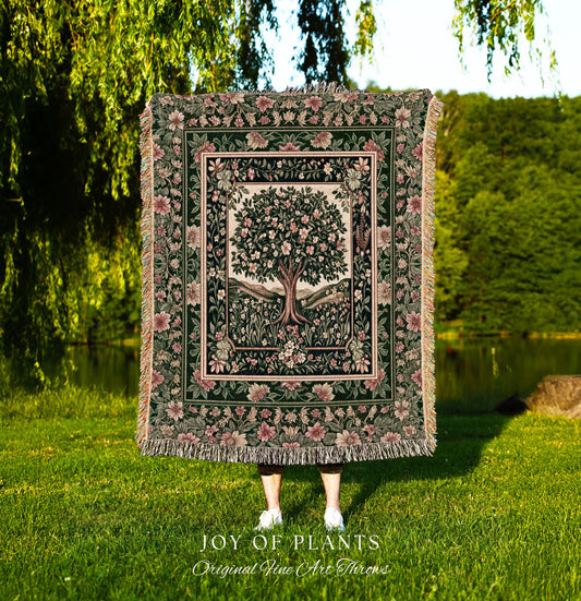 May include: A tapestry throw blanket with a floral and tree design, held by a person in a grassy field. The blanket features a central tree image surrounded by flowers and a dark green border. The text "JOY OF PLANTS" and "Original Fine Art Throws" is visible.