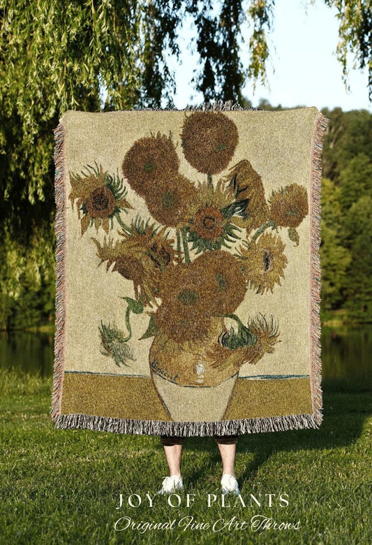 May include: A woven throw blanket featuring a detailed depiction of Van Gogh's "Sunflowers" painting. The artwork showcases sunflowers in a vase, rendered in shades of brown, yellow, and green. The blanket has a fringed edge and the text "JOY OF PLANTS" and "Original Fine Art Throws".