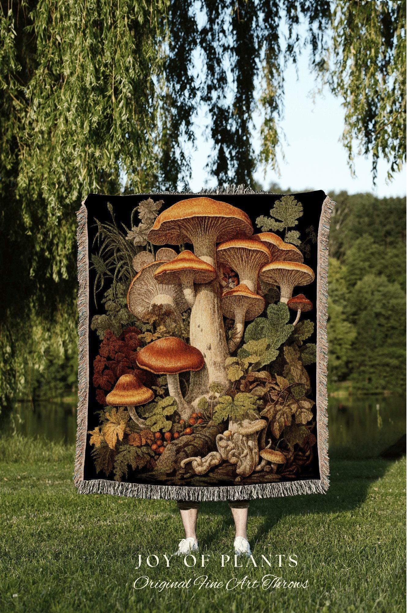May include: A black throw blanket featuring a detailed illustration of various mushrooms in shades of brown, orange, and cream, surrounded by green foliage. The blanket has a fringed edge and the text "JOY OF PLANTS Original Fine Art Throws".
