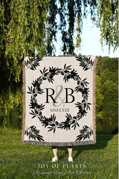 May include: A beige throw blanket with a black olive branch wreath design. The center features the letters "R & B" and the Roman numeral "MMXXIII". The blanket has a fringed edge and the text "JOY OF PLANTS" at the bottom.