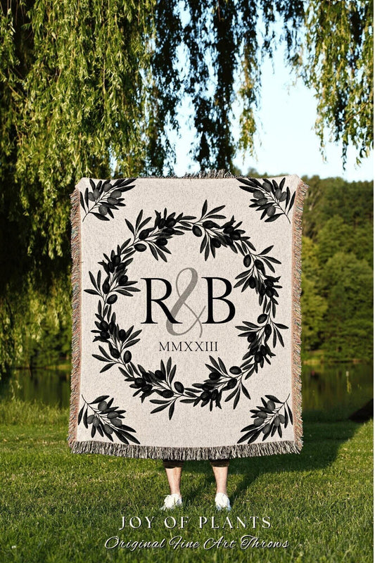 May include: A beige throw blanket with a black olive branch wreath design. The center features the letters "R & B" and the Roman numeral "MMXXIII". The blanket has a fringed edge and the text "JOY OF PLANTS" at the bottom.