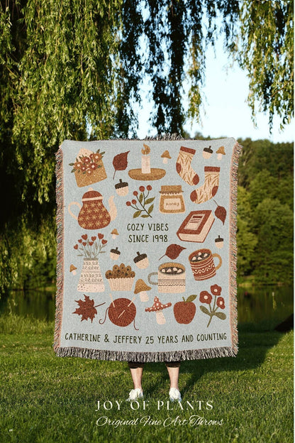 May include: A light blue throw blanket with a repeating pattern of cozy autumn-themed illustrations. The design includes a teapot, candles, socks, and the text "COZY VIBES SINCE 1998". The bottom of the blanket reads "CATHERINE & JEFFERY 25 YEARS AND COUNTING".