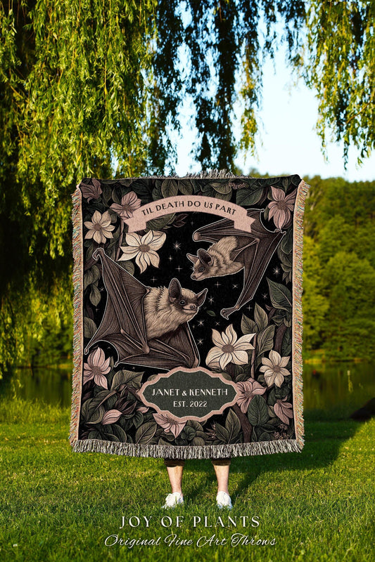 May include: A black throw blanket featuring a bat design with floral accents and the text "TIL DEATH DO US PART." The blanket includes the names "JANET & KENNETH" and the year "2022." The bottom of the blanket has the text "JOY OF PLANTS Original Fine Art Throws."