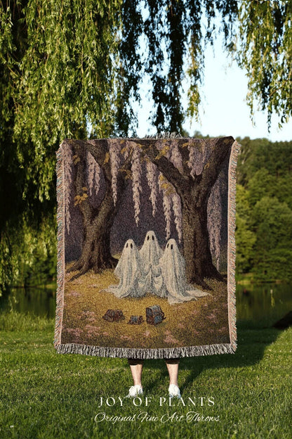 May include: A woven throw blanket featuring a forest scene with three ghost figures. The blanket has a fringed edge and the text "JOY OF PLANTS Original Fine Art Throws" at the bottom. The color palette includes shades of brown, gray, and white.
