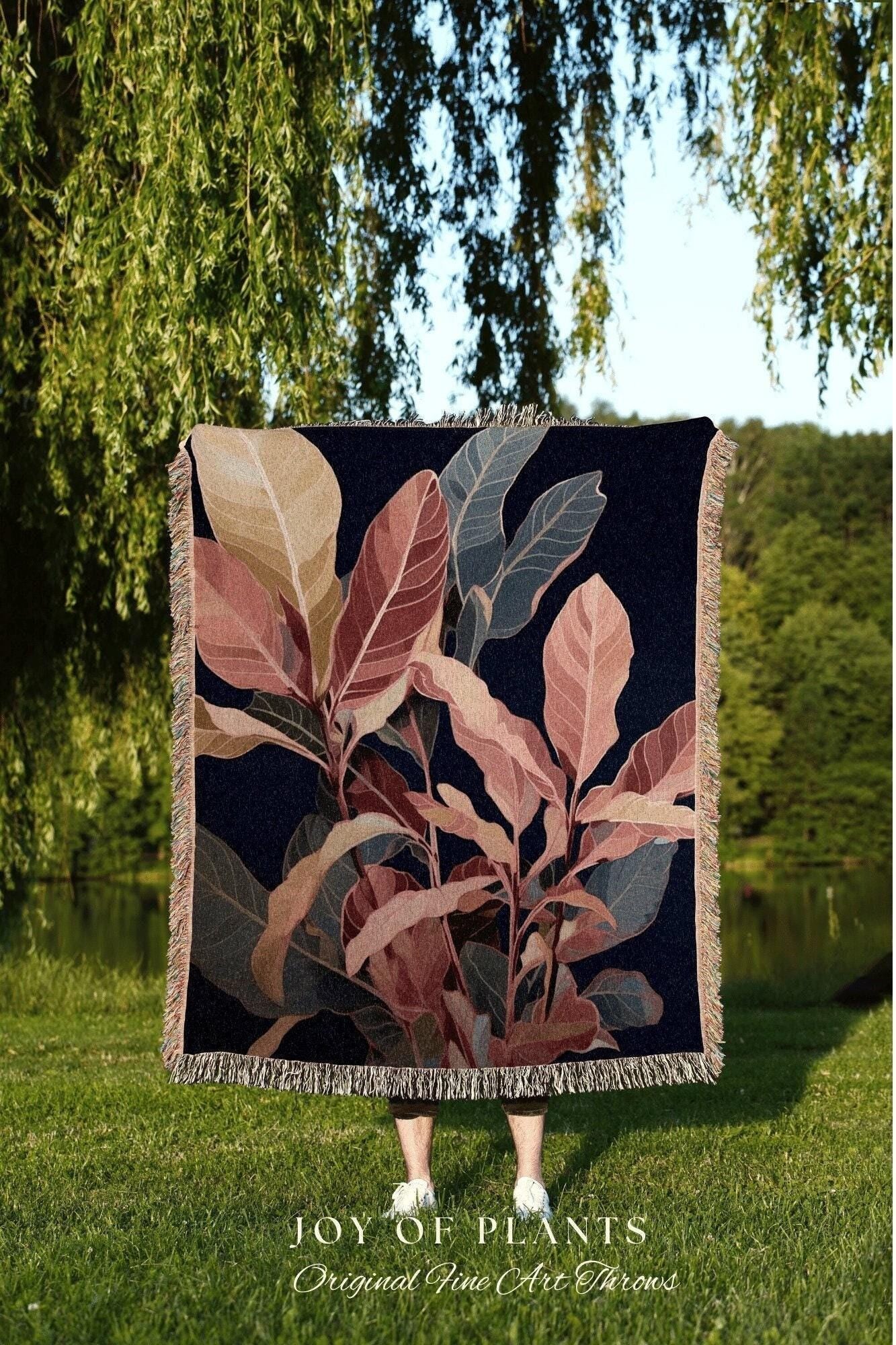 May include: A dark blue throw blanket with a botanical design featuring large leaves in shades of pink, beige, and gray. The blanket has a fringed edge and the text "JOY OF PLANTS Original Fine Art Throws" at the bottom.