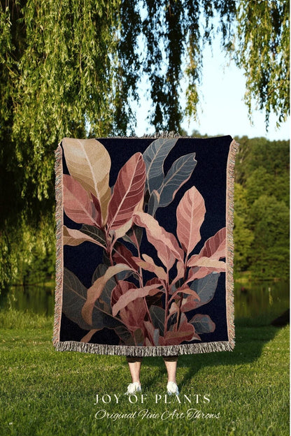 May include: A dark blue throw blanket with a botanical design featuring large leaves in shades of pink, beige, and gray. The blanket has a fringed edge and the text "JOY OF PLANTS Original Fine Art Throws" at the bottom.