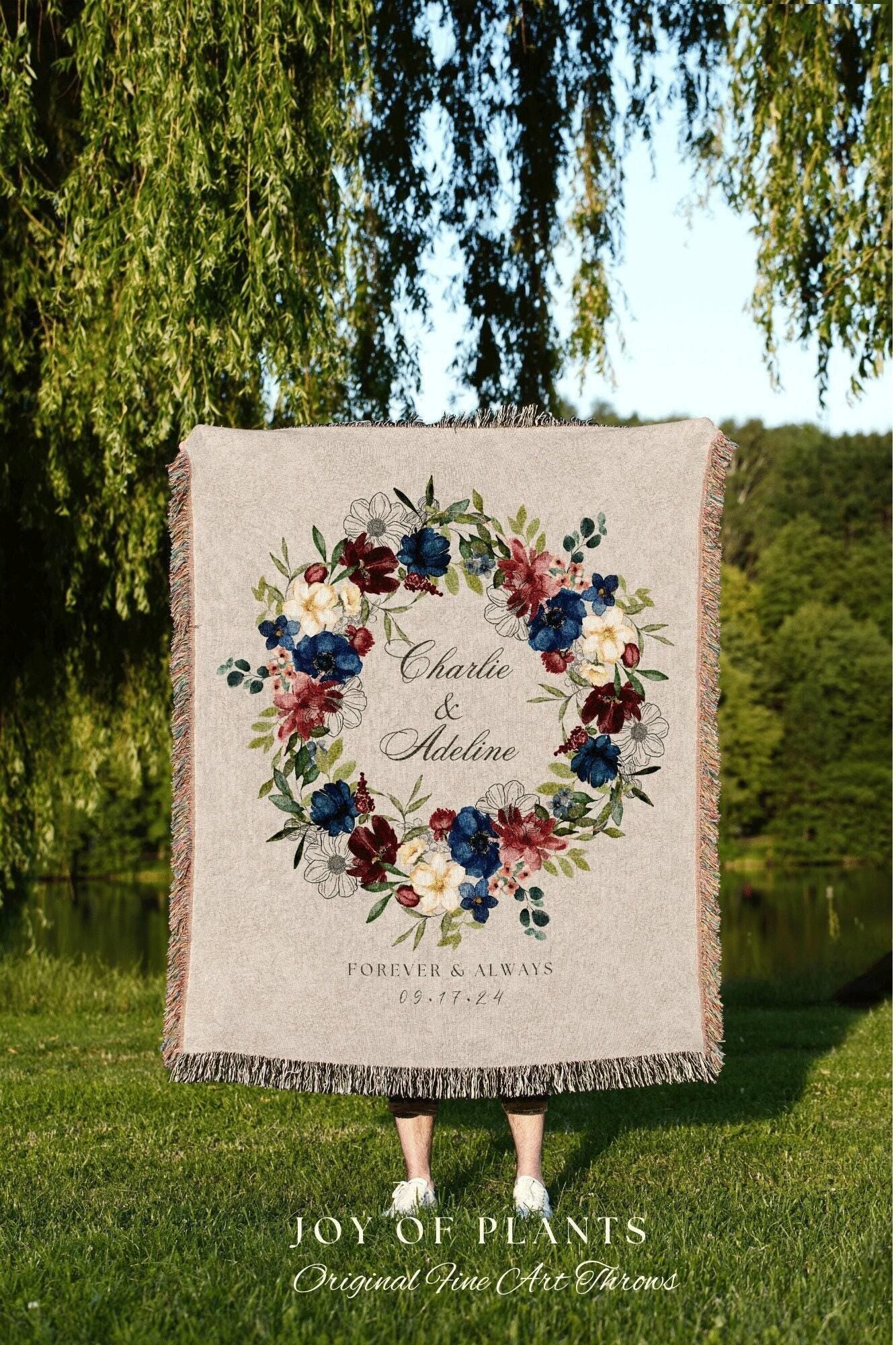 May include: A beige throw blanket with a floral wreath design in burgundy, navy, and cream colors. The text "Charlie & Adeline" and "Forever & Always 09.17.24" are displayed within the wreath. The blanket has a fringed edge.