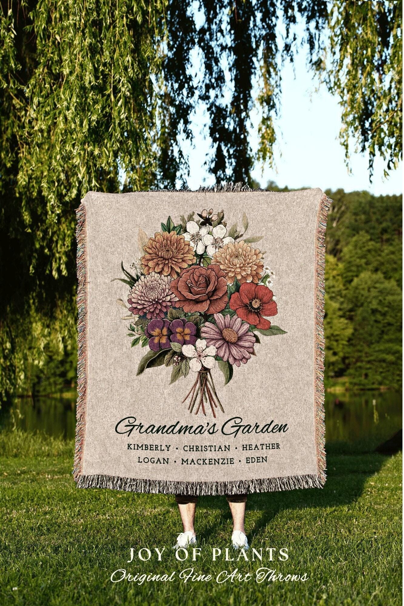 May include: A woven throw blanket with a floral design. The blanket features a bouquet of flowers in shades of red, orange, pink, purple, and white. The text 'Grandma's Garden' is printed below the bouquet, followed by a list of names: Kimberly, Christian, Heather, Logan, Mackenzie, Eden. The text 'JOY OF PLANTS Original Fine Art Throws' is printed below the list of names.