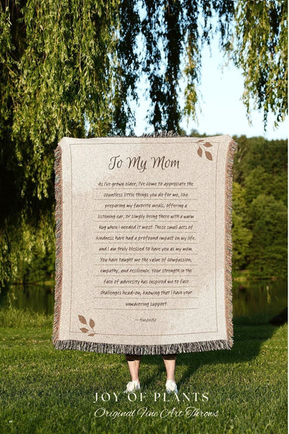 May include: A beige throw blanket with a printed message "To My Mom" and a heartfelt message. The blanket has a fringed edge and is held outdoors in front of a green background. The Joy of Plants logo is at the bottom.