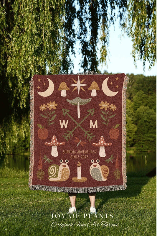 May include: A burgundy throw blanket with a whimsical forest design. It features mushrooms, moons, stars, and snails. The blanket has a fringed edge and the text "SHARING ADVENTURES SINCE 2023". The words "W" and "M" are also included.
