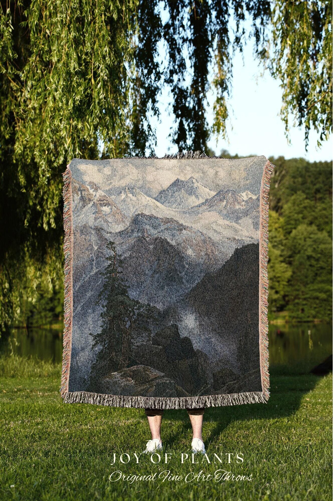 May include: A woven throw blanket featuring a mountain landscape scene in shades of gray, blue, and black. The blanket has a fringed edge and the text "JOY OF PLANTS Original Fine Art Throws" at the bottom.