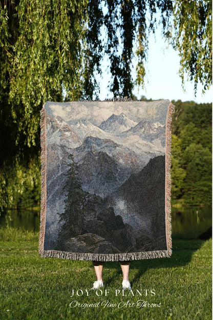 May include: A woven throw blanket featuring a mountain landscape scene in shades of gray, blue, and black. The blanket has a fringed edge and the text "JOY OF PLANTS Original Fine Art Throws" at the bottom.