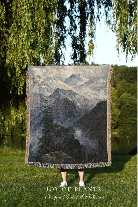 May include: A woven throw blanket featuring a mountain landscape scene in shades of gray, blue, and black. The blanket has a fringed edge and the text "JOY OF PLANTS Original Fine Art Throws" at the bottom.