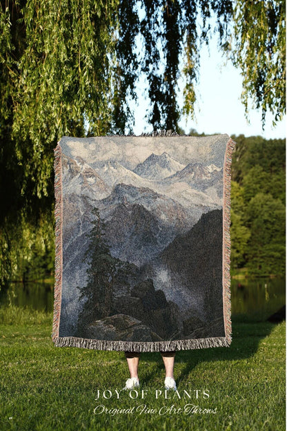 May include: A woven throw blanket featuring a mountain landscape scene in shades of gray, blue, and black. The blanket has a fringed edge and the text "JOY OF PLANTS Original Fine Art Throws" at the bottom.