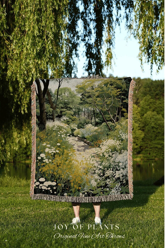 May include: A woven throw blanket featuring a detailed landscape scene with a path, trees, and wildflowers in shades of green, yellow, and white. The blanket has a fringed edge and the text "JOY OF PLANTS Original Fine Art Throws" at the bottom.