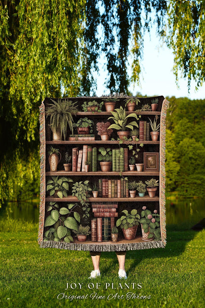 May include: A woven throw blanket featuring a detailed illustration of a bookshelf filled with potted plants and books. The blanket has a dark background and a fringed edge. The text "JOY OF PLANTS Original Fine Art Throws" is at the bottom.