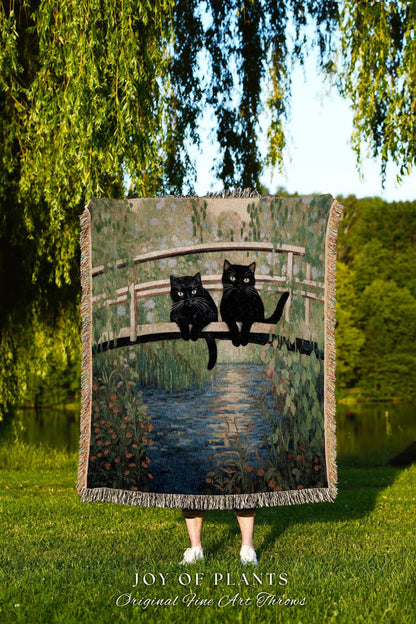 May include: A woven throw blanket featuring a painting of two black cats sitting on a bridge over a river. The artwork has a soft, impressionistic style with green and blue tones. The blanket has a fringed edge and the text "JOY OF PLANTS Original Fine Art Throws".