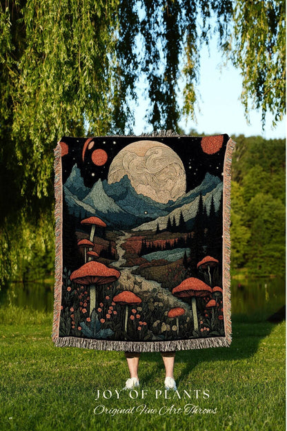 May include: A woven throw blanket featuring a landscape design with large orange mushrooms, mountains, and a large moon. The blanket has a black background and fringed edges. The text "JOY OF PLANTS Original Fine Art Throws" is at the bottom.